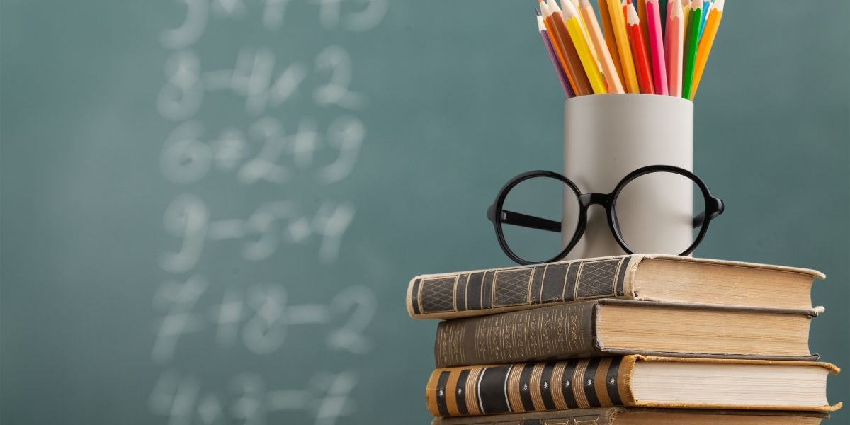 Stos książek, okulary i kubek z kredkami na tle tablicy szkolnej